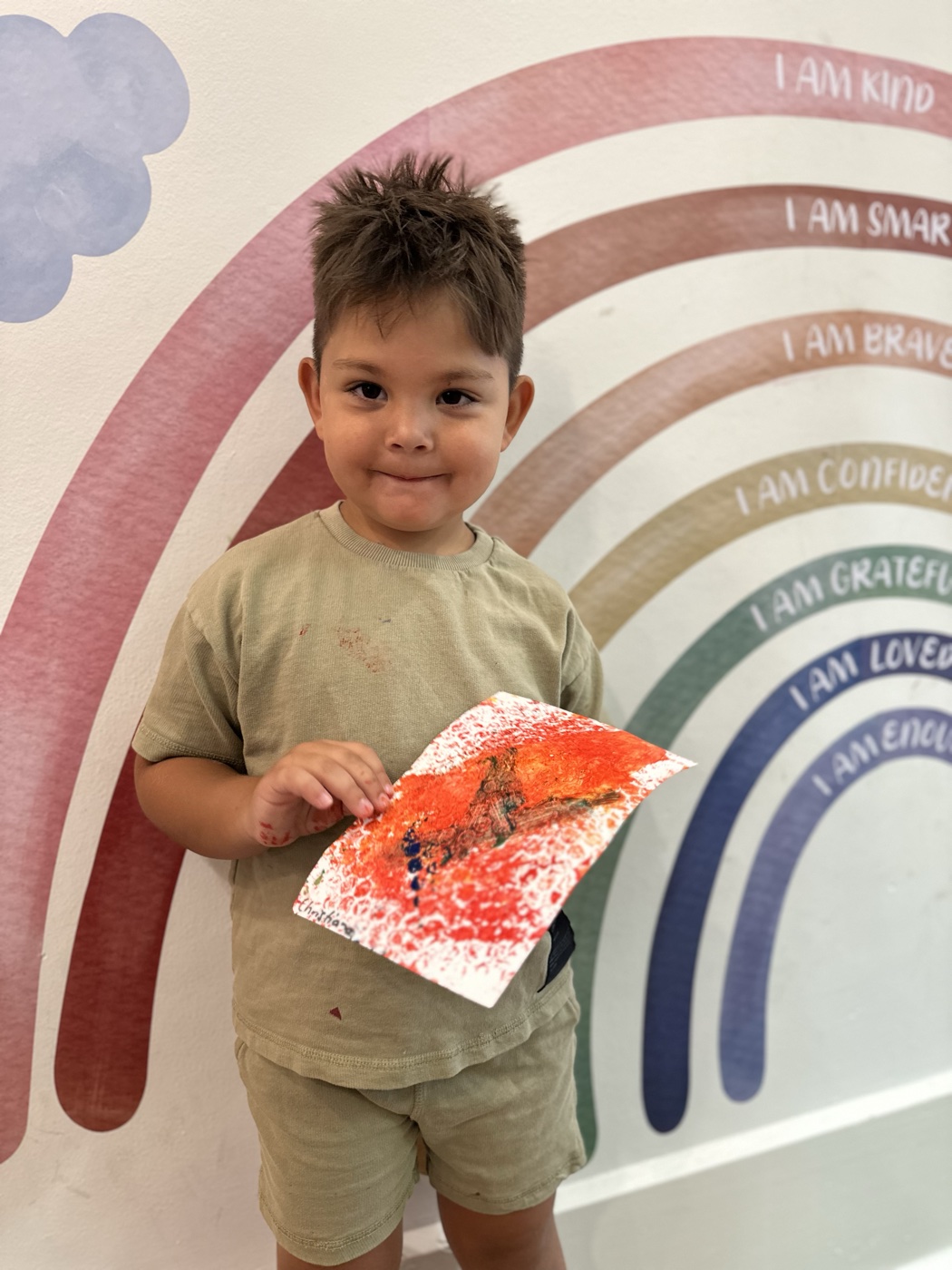 Child holding their painting in front of the Mini Ivy affirmation rainbow: I am kind, I am smart, I am brave, I am confident