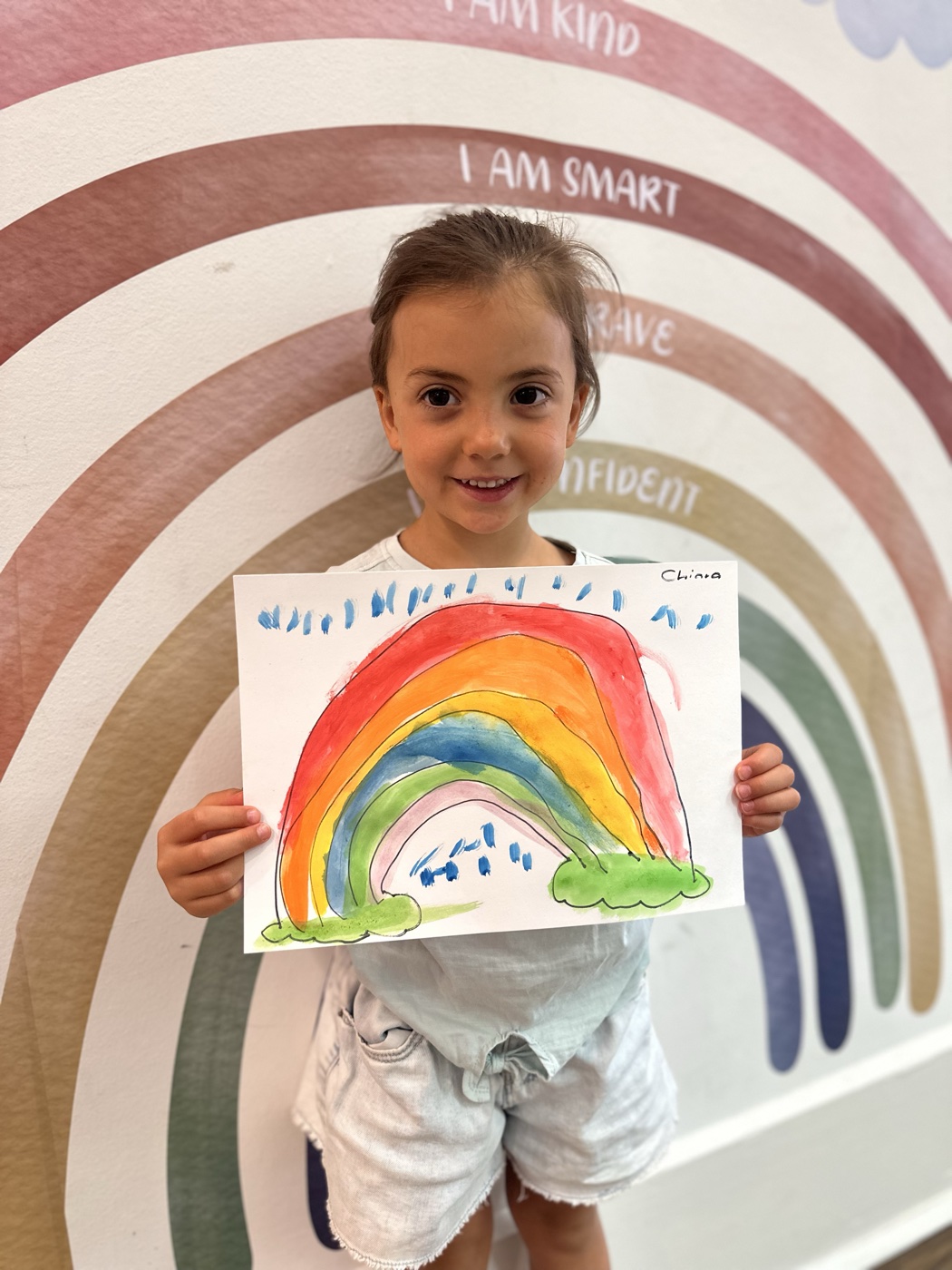 Child holding her rainbow painting in front of the Mini Ivy affirmation wall