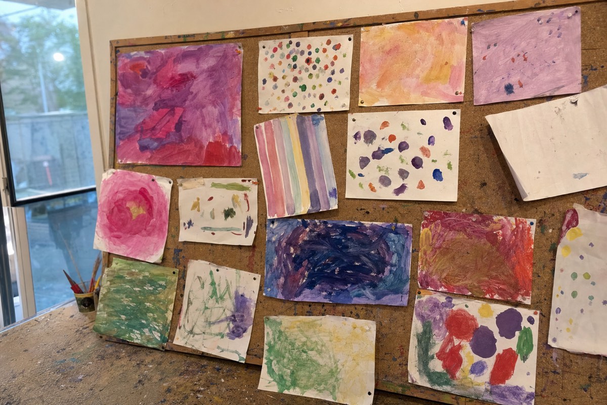 A wall of young children's paintings pinned up unevenly, none alike: stripes, dots, washes and scribbles